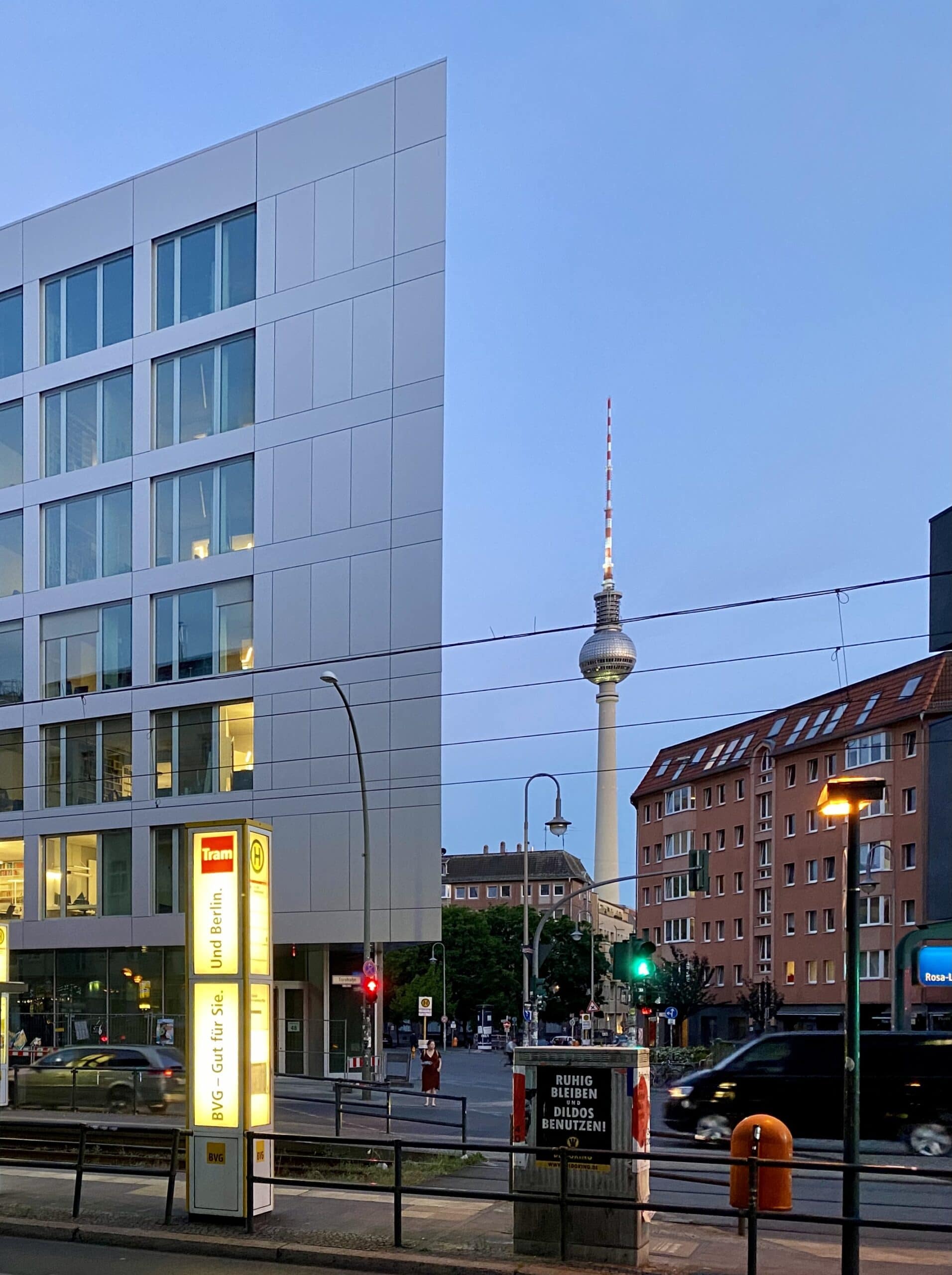 Moderne Gebäude bei Dämmerung, Berliner Fernsehturm sichtbar, beleuchtete Straße, ruhige Stimmung.